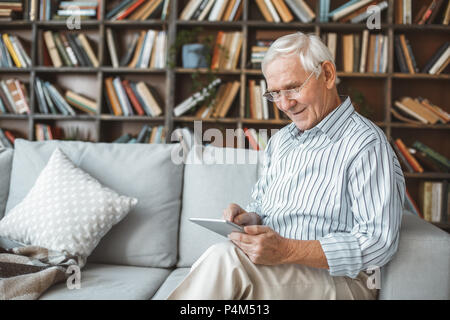 Aged male at home in the living room sitting on the coach using digital tablet smiling Stock Photo