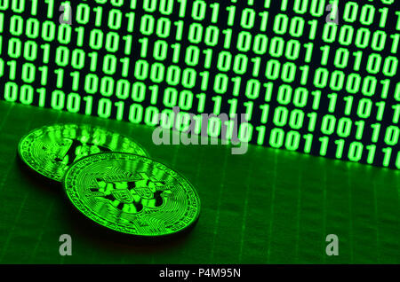 A pair of bitcoins lies on a cardboard surface on the background of a monitor depicting a binary code of bright green zeros and one units on a black b Stock Photo