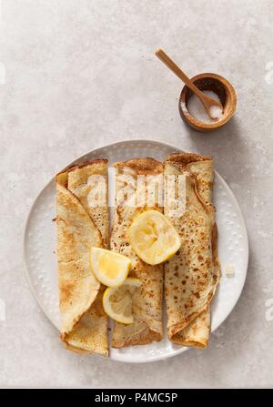 three rolled pancakes pn a off white texture plate with lemon wedges on a stone surface propped with wooden sugar bowl and spoon Stock Photo