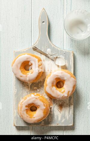 Three iced doughnuts on a grey wooden chopping board with a spoon Stock Photo