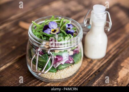 A quinoa salad with lambs lettuce, radicchio, rocket, croutons, goat's cheese and horned violets in a glass jar, with dressing in a glass bottle Stock Photo