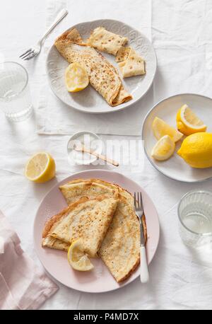 Two plates of folded and rolled pancakes served with lemon wedges on white linen covered table and water glasses Stock Photo