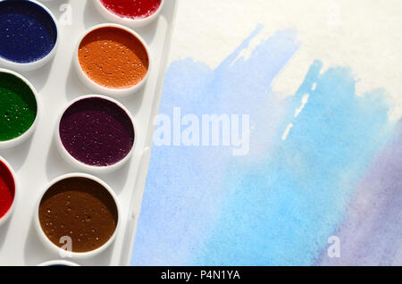 A new set of watercolors lies on a sheet of paper, which shows an abstract watercolor drawing in the form of blue strokes. The concept of amateur pain Stock Photo