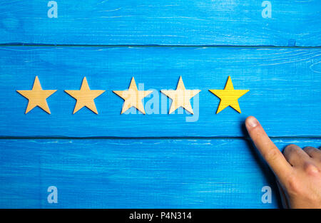The male hand points to the fifth yellow star on a blue wooden background. Five Stars. Rating of restaurant or hotel, application. Evaluation of quali Stock Photo