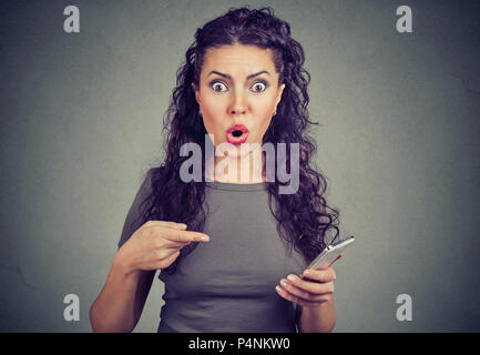 young woman looking with terrified expression at camera pointing at smart phone, after reading shocking news on webpage isolated on gray background. Stock Photo