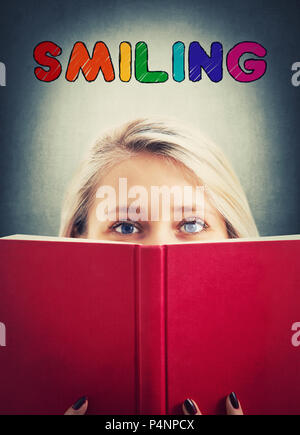 Close up portrait of pretty student girl hiding her personality behind a red opened book like a mask and colorful smiling text inscription above her h Stock Photo