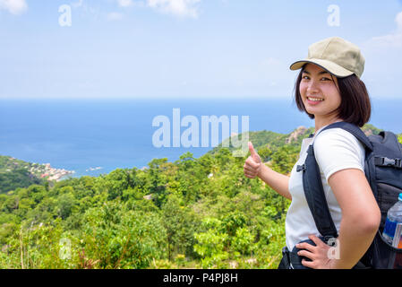 Women tourist with a backpack wear cap raise thumbs up for the beautiful nature landscape blue sea and sky from high scenic viewpoint at Koh Tao, Sura Stock Photo