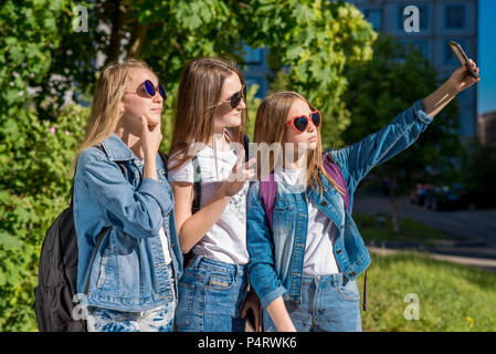 Girls schoolgirls in summer in park. He takes pictures of himself on the phone. Happy smiling pose on camera. In denim clothes. Female students wearing sunglasses. Outdoors in the city. Stock Photo