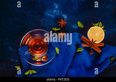 Hot drink header with glass teacup, linen napkin, lemon slices, cinnamon, anise stars and mint leaves on a dark background. Dark food photography flat Stock Photo