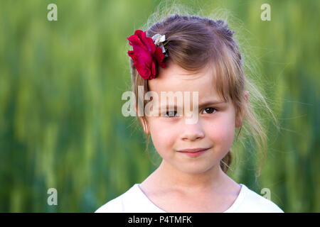 Portrait of beautiful small preschool blond girl with nice gray eyes and red rose in hair smiling dreamily in camera on blurred bright green backgroun Stock Photo