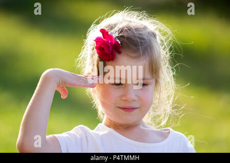 Portrait of cute small funny blond girl with nice gray eyes and red rose in hair with right arm raised in salute gesture on blurred bright green backg Stock Photo