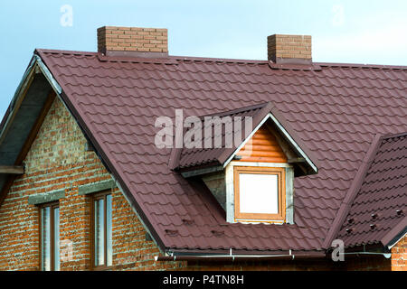 Close-up detail of new brick house top with brown shingle roof, two brick chimneys and plastic attic windows on bright blue sky background. Real estat Stock Photo