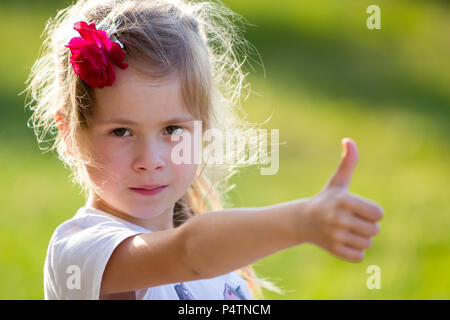 Portrait of cute small blond serious girl in white T-shirt with red rose in hair showing thumb-up gesture to camera on blurred bright green summer bac Stock Photo