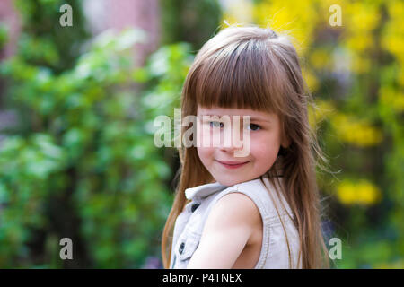 Portrait of a pretty little long-haired blond preschool girl in sleeveless white dress smiling shyly into camera against blurred outdoors background.  Stock Photo