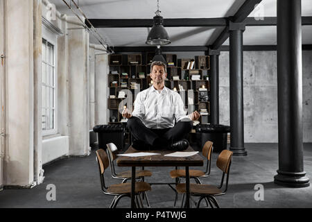 Portrait of mature businessman doing yoga on desk in loft office Stock Photo