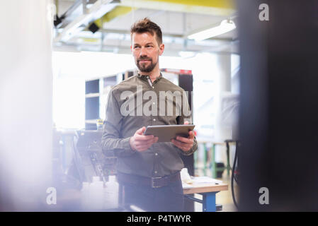 Smiling man standing in factory with tablet looking sideways Stock Photo