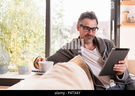 Portrait of mature man with cup of coffee sitting on couch using tablet Stock Photo