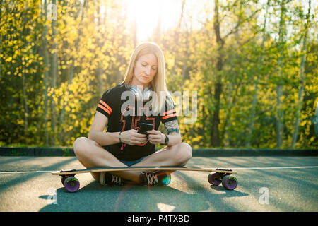 Blond woman with longboard sitting on street using smartphone Stock Photo