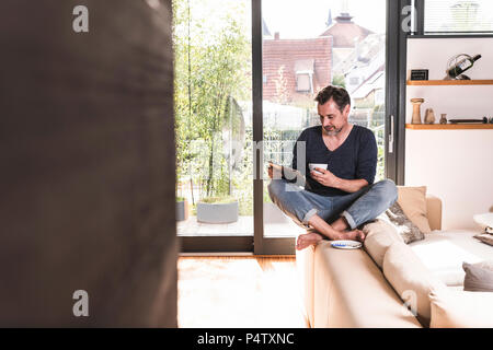Mature man with cup of coffee sitting on backrest of couch using tablet Stock Photo