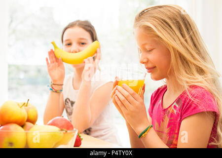 Portrait of girl drinking glass of orange juice while her friend having fun with banana Stock Photo