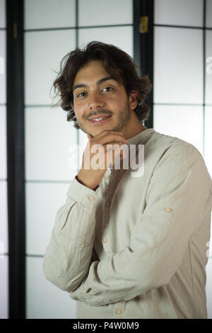 An handsome young man, 20Y, is posing in studio and touching his chin with satisfaction. White skirt. Retroilluminated booths in the background. Stock Photo