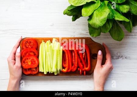 A girl is holding a board with freshly sliced vegetables: tomato, cucumber and pepper. Washed fresh bunch of spinach leaves . Top view. Stock Photo