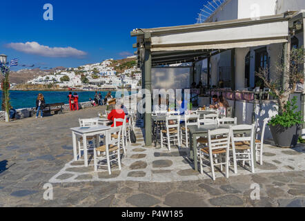 Mykonos, Greece crowd at tavern style fast food restaurant. People eating on outdoor seating white tables by town waterfront in central Manto Square. Stock Photo
