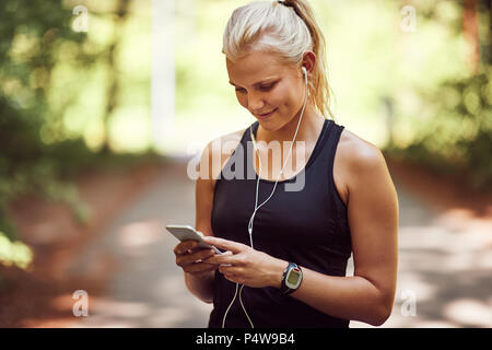 Smiling young blonde woman in sportswear queueing up a music playlist on her cellphone before a run through a forest Stock Photo