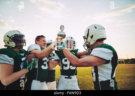 Ecstatic group of American football players standing in a huddle on a field raising a championship trophy in celebration Stock Photo