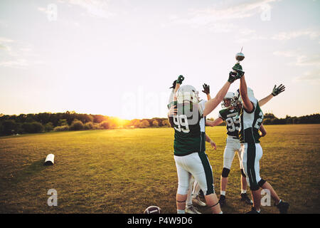 Excited group of American football players standing together in a huddle and raising a championship trophy in celebration Stock Photo