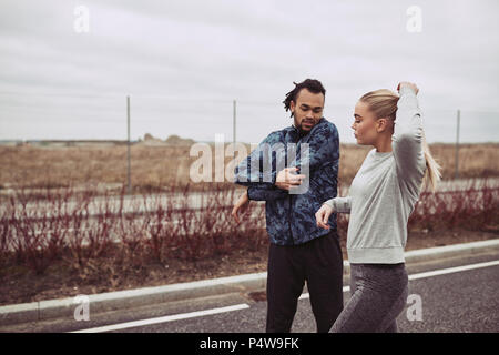 Diverse young couple doing stretches before going for a run along a road in the country on an overcast day Stock Photo