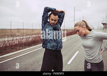 Young couple in sportswear doing stretches before going for a run together along a country road on an overcast day Stock Photo