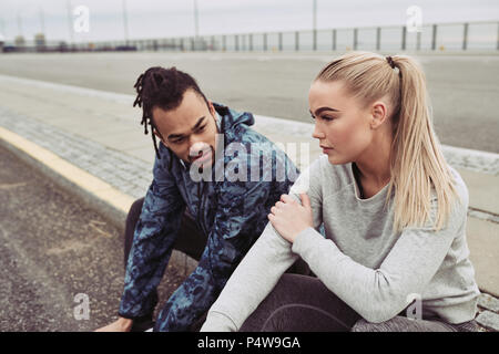 Diverse young couple in sportswear sitting on the side of a road taking a break from a run on an overcast day Stock Photo