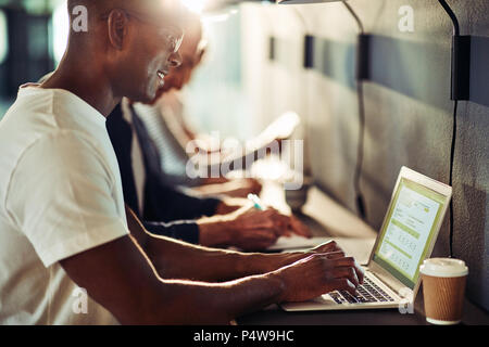 Smiling young African designer working on a laptop while sitting with colleagues at a table in an office Stock Photo