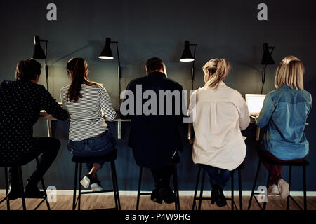 Rearview of a group of businesspeople working together while sitting in a row at a table in a dark office Stock Photo