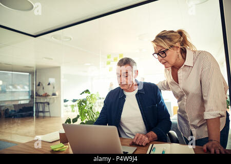 Two focused mature work colleagues discussing a project together over a laptop at a desk in a modern office Stock Photo