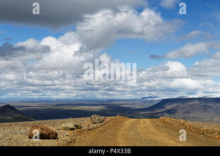 A gravel road leads over a small hill, behind it is a wide landscape with plain, mountain ranges and glaciers to see, above blue sky with white and gr Stock Photo