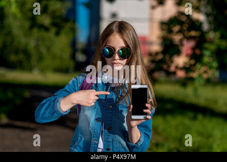 Little girl schoolgirl in sunglasses. Summer in nature. In his hands holds a smartphone. A finger points to phone. The concept is a new gadget app. Emotion focus concentration. Stock Photo