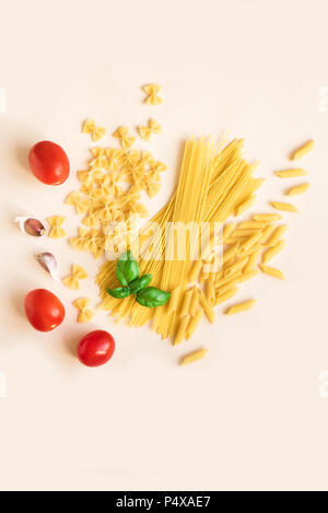 Various uncooked raw pasta with basil,tomatoes and garlic on pink pastel background. Italian pasta: penne, farfalle, spaghetti. Stock Photo