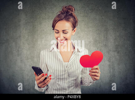 Happy cute woman looking at her cellphone holding a red heart shape Stock Photo