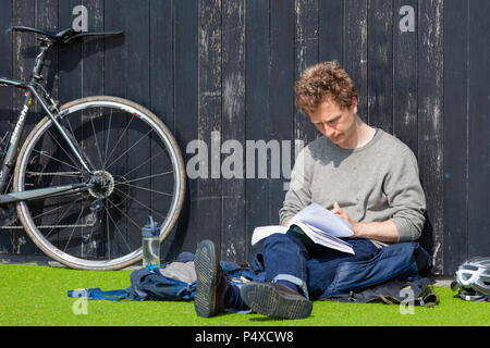 A man reads a book sitting on fake green grass with a bike propped up against a timber wall Stock Photo