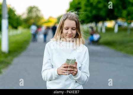 A teenage girl. In the summer in city. In his hands holds a smartphone. Listens to music watching video on the Internet. Writes messages on social networks. Smiles happily. Stock Photo