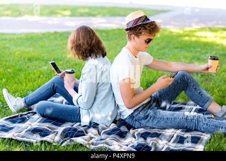 A guy with a girl in summer relax in nature. He holds a smartphone in his hands and reads correspondence. He holds a glass of coffee or tea in his hands. Enjoys relaxing on a plaid. Stock Photo