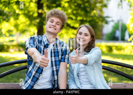 Young couple girl and boy. Summer in park on a bench. Smiles happily. Hard hands showing thumbs up, like. Emotionally gestures. The concept of success and success. Stock Photo