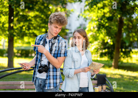 The guy with the girl is young tourists. In summer in park in nature. In the hands holds a smartphone road map. On the neck is a digital camera. Gestures of the hands show the direction of movement. Stock Photo