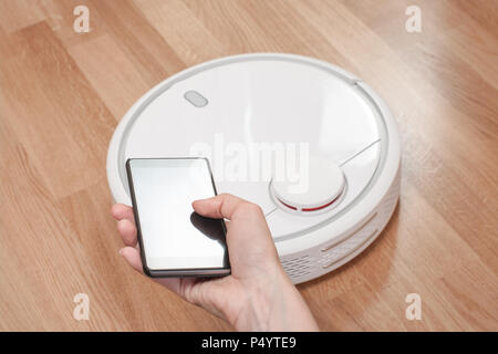 womens hand using mobile to control white robotic vacuum cleaner. modern smart cleaning technology. Stock Photo