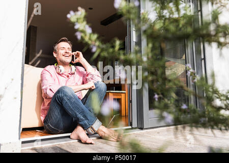 Laughing mature man on the phone sitting at open terrace door Stock Photo
