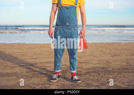 Man in dungarees standing on the beach holding bottles of soft drinks Stock Photo