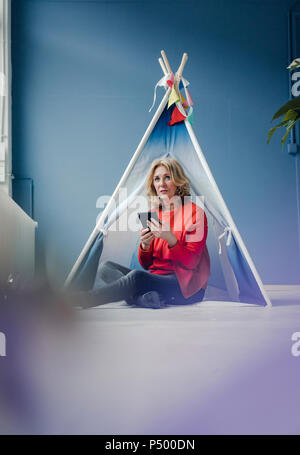 Senior woman sitting at teepee indoors holding phablet Stock Photo