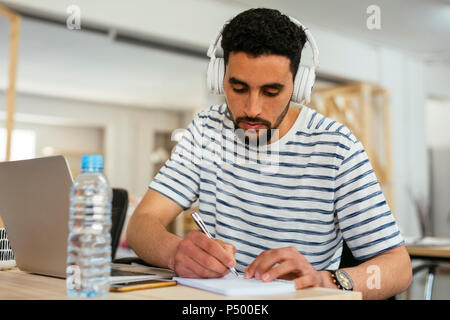 Young man wearing headphones taking notes at desk in office Stock Photo